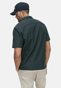 Mörkgrön kortärmad polotröja med krage, texturerat tyg och rak fåll. Kombineras med ljusa khakibyxor och en marinblå keps.