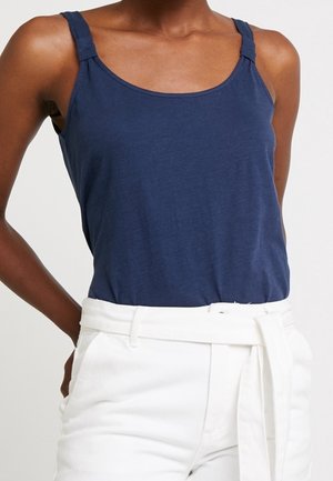 Granatowy top na ramiączkach z okrągłym dekoltem i szerokimi ramionkami. Wykonany z miękkiego materiału, o luźnym kroju, zestawiony z białymi spodniami z paskiem.