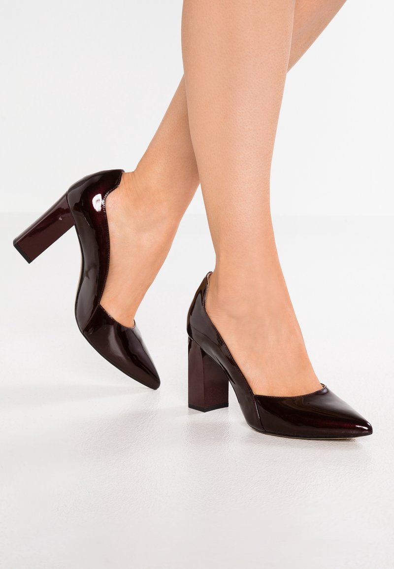 Jambes portant des chaussures à talons hauts bordeaux foncé brillants avec de larges talons blocs sur un sol blanc.