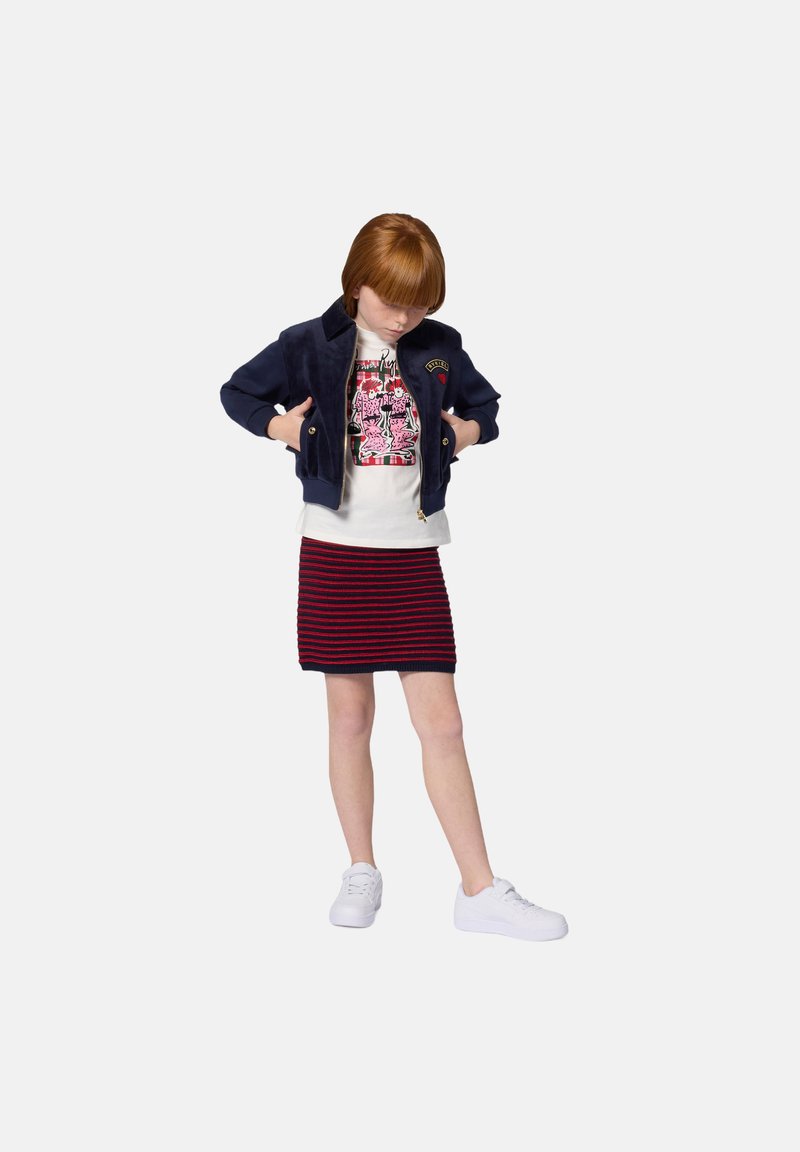 Child wearing a navy velour jacket, white graphic tee, and a red and navy striped skirt, paired with white sneakers.