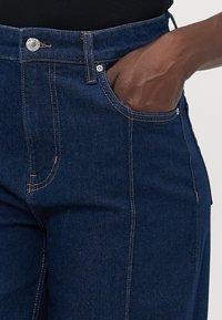 Gros plan sur une main reposant à l'intérieur de la poche avant d'un jean en denim bleu foncé avec des coutures visibles et un bouton de fermeture.