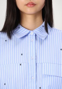 Chemise à rayures bleues avec de petits imprimés noirs en forme de "E", col classique, fermeture par boutons et deux poches poitrine. Tissu doux avec une finition nette.