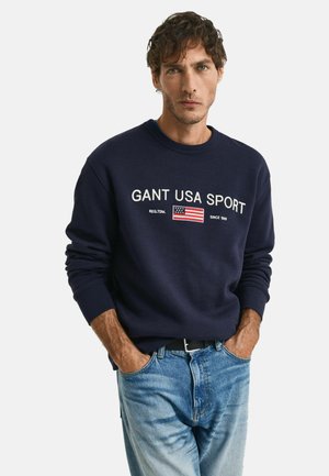 Navy blue sweatshirt made of soft fabric, featuring white text "GANT USA SPORT" and a small USA flag graphic on the chest.