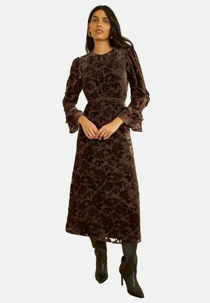 Langes braunes Kleid mit floralem Muster aus strukturiertem Stoff. Verfügt über Puffärmel und taillierten Schnitt, kombiniert mit schwarzen Stiefeln mit hohem Absatz.