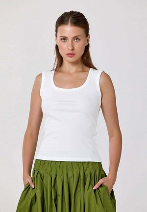 Jeune femme aux cheveux bruns portant un haut blanc sans manches et une jupe plissée verte, debout les mains dans les poches devant un fond uni.