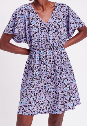 Femme portant une courte robe fleurie violette avec des manches flottantes et une taille froncée, debout les mains sur les hanches.