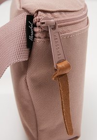 Gros plan d’un sac en tissu Herschel rose poussiéreux avec fermeture éclair et tirette en cuir sur fond blanc.