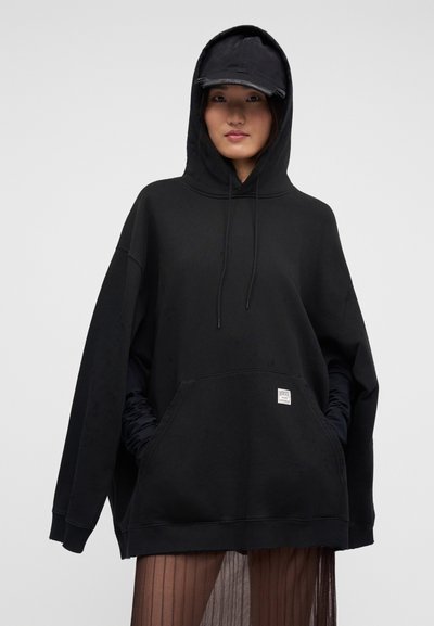 Νεαρό άτομο που φοράει μαύρο φαρδύ φούτερ με τσέπη μπροστά, μαύρο καπέλο και διαφανή πλισέ φούστα, στέκεται μπροστά σε απλό φόντο.