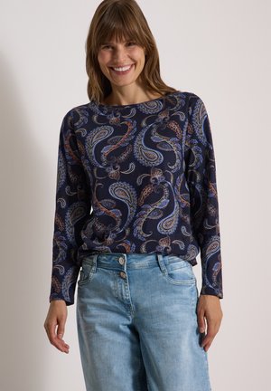 Femme souriante portant un haut bleu marine à manches longues avec un motif cachemire et un jean en denim bleu clair sur un fond uni.