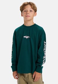 Lange mouwen, donkergroen katoenen shirt met witte logo op de borst en gebrandmerkte tekst langs de mouwen. Zachte stof met een relaxte fit.