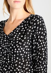 Blouse noire avec un col en V, présentant des motifs d'étoiles blanches. Tissu léger à surface texturée et coupe décontractée. Manches longues.
