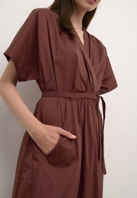 Brown, short-sleeve wrap dress made of smooth fabric, featuring a tie belt and side pocket, with relaxed fit and soft texture.