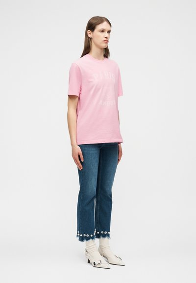 Γυναίκα που στέκεται φορώντας ένα ανοιχτό ροζ κοντομάνικο πουκάμισο, τζιν μπλε με διακοσμητικά λουλούδια στους ξεφτισμένους ποδόγυρους, λευκές κάλτσες και μυτερές παπούτσια.