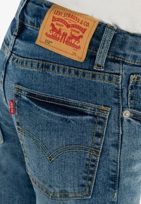 Jeans de denim azul con una etiqueta de cuero marrón que presenta dos caballos y "LEVI STRAUSS & CO." El bolsillo trasero tiene una etiqueta de logo roja y costuras amarillas.