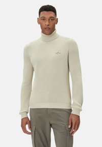 Maglione a collo alto color beige chiaro con motivo a maglia lavorata a mano, polsini e orlo a coste, e piccolo logo ricamato sul petto. Indossato con pantaloni grigi.