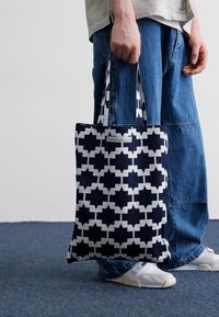 Tamnoplava torba s bijelim geometrijskim uzorkom, izrađena od čvrstog materijala. Ima dva remena za rame i jednostavan pravokutni oblik.