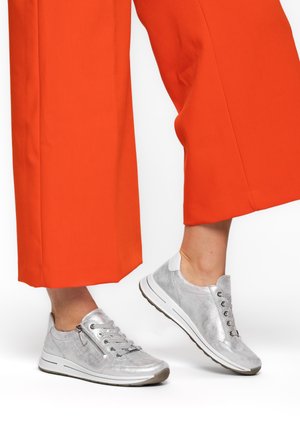 Silver sneakers with side zipper and laces worn with bright orange wide-leg cropped pants, shown mid-step against white background.