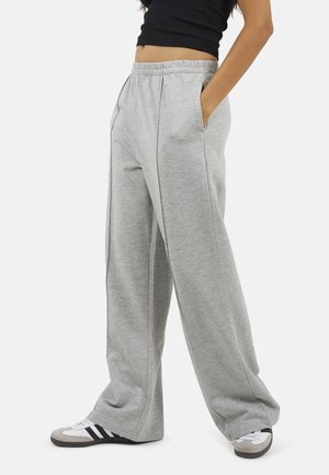 Persoon draagt losse lichtgrijze joggingbroek, zwarte cropped top en witte sneakers met zwarte strepen, staat met één hand in de zak.