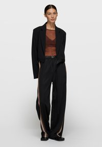 Blazer negro, top marrón de malla, pantalones negros con franjas laterales blancas y marrones. Texturas suaves, corte entallado, detalles de diseño mínimos.