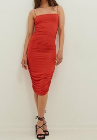 Rotes, tailliertes Midi-Kleid mit dünnen Trägern und Raffung an den Seiten. Kombiniert mit schwarzen, schmalen, hochhackigen Sandalen.