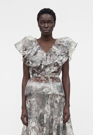 Γυναίκα με διάφανο φόρεμα με floral σχέδιο, μεγάλα βολάν στους ώμους και στρωτή φούστα, στέκεται μπροστά σε απλό ανοιχτό φόντο.