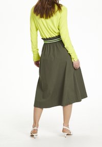 Falda midi verde con cintura fruncida y banda a rayas en contraste. Combinada con una blusa ajustada de manga larga amarilla brillante y tacones blancos.