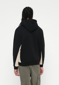 Sudadera negra con capucha, paneles laterales beige en contraste y puños acanalados. Material de algodón con un corte relajado y detalles mínimos.
