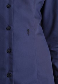 Dunkelblauses Hemd aus glattem Stoff mit schwarzen Knöpfen und einem kleinen schwarzen Rosen-Stickereibezug auf der linken Seite.