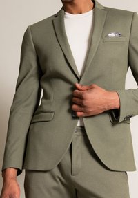 Blazer su misura verde oliva con rever a occhiello, due tasche frontali e un fazzoletto da taschino grigio chiaro. Indossato sopra una camicia bianca. Tessuto texture.