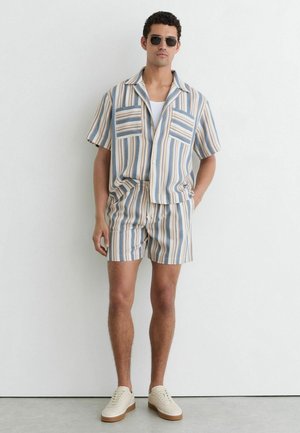 Mann mit Sonnenbrille, gestreiftem Kurzarmhemd und passenden Shorts, der mit den Händen in den Taschen an eine weiße Wand lehnt.