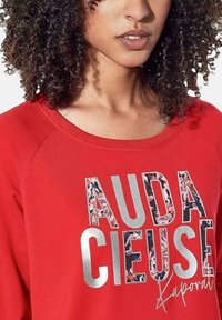Rode sweatshirt met een ronde hals en raglanmouwen. Heeft grote, multicolore grafische tekst en zilveren accenten. Zachte stofstructuur.