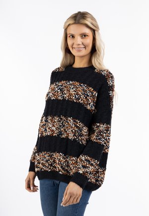 Giovane donna con lunghi capelli biondi indossa un maglione nero con righe testurizzate marrone, bianco e blu navy e jeans blu, sorridendo dolcemente.