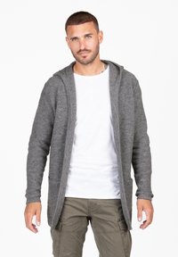 Cardigan grigio a maglia con cappuccio, due tasche frontali e maniche lunghe, indossato sopra una t-shirt bianca. Tessuto texturizzato e design frontale aperto.