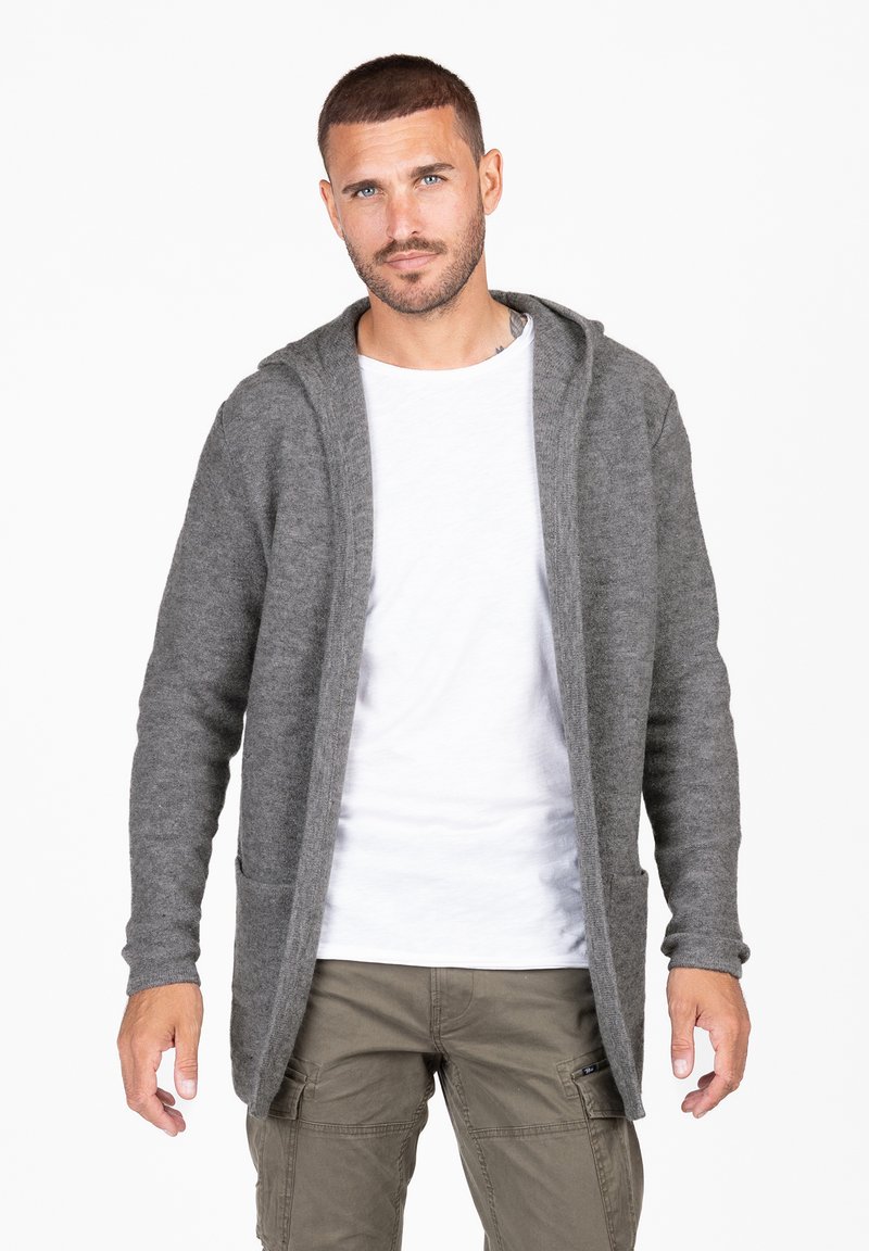 Cardigan gris de punto con capucha, dos bolsillos frontales y mangas largas, usado sobre una camiseta blanca. Tejido texturizado y diseño de apertura frontal.