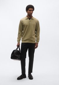 Olíva zöld pulóver gallérral, fekete nadrág és fekete texturált kézitáska. Férfi áll sötét cipőben fehér háttér előtt.