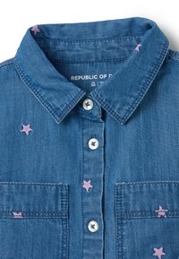 Camisa de mezclilla de tela azul claro, diseño de botones, cuello y bordado de estrellas rosa. Cuenta con dos bolsillos en el pecho y botones blancos.