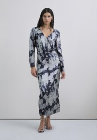 Nainen, joka käyttää pitkähihaista, hopea- ja mustat vivahtavaa midimekkoa, jossa on syvä V-pääntie, sekä hopeita niittikorkokenkiä, seisoo harmaalla lattialla.