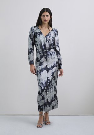 Mujer con un vestido midi de manga larga, en tonos plateados y negros con estampado tie-dye, con un escote en V profundo y sandalias altas de tiras plateadas, de pie sobre un suelo gris.