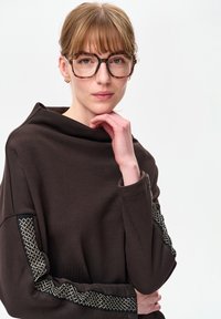 Brązowy, miękki top z dekoltem typu wodospad, ozdobiony teksturowanymi czarno-złotymi akcentami na rękawach. Modelka w okularach, z ręką na podbródku.