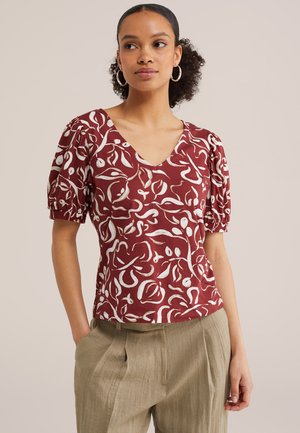 Frau trägt eine rote V-Ausschnitt-Bluse mit weißen abstrakten Blumenmustern und Puffärmeln, kombiniert mit beigen Hosen mit hoher Taille.