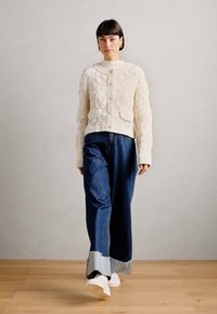 Cremfarbener Strickcardigan mit strukturiertem Blumenmuster und Knopfleiste; kombiniert mit weit geschnittenen blauen Jeans aus Denim mit umgeschlagenen Säumen.