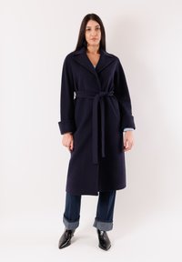 Donna che indossa un lungo cappotto blu scuro con cintura, jeans scuri risvoltati e stivali neri a punta, in piedi davanti a uno sfondo chiaro e uniforme.