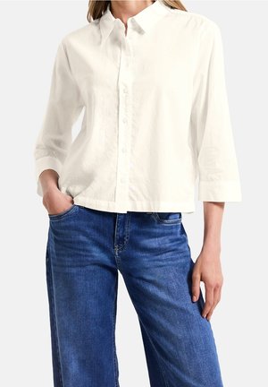 Frau trägt ein weißes, langärmliges Hemd mit Knöpfen und blaue Jeans, eine Hand in der Tasche und die andere entspannt an der Seite.