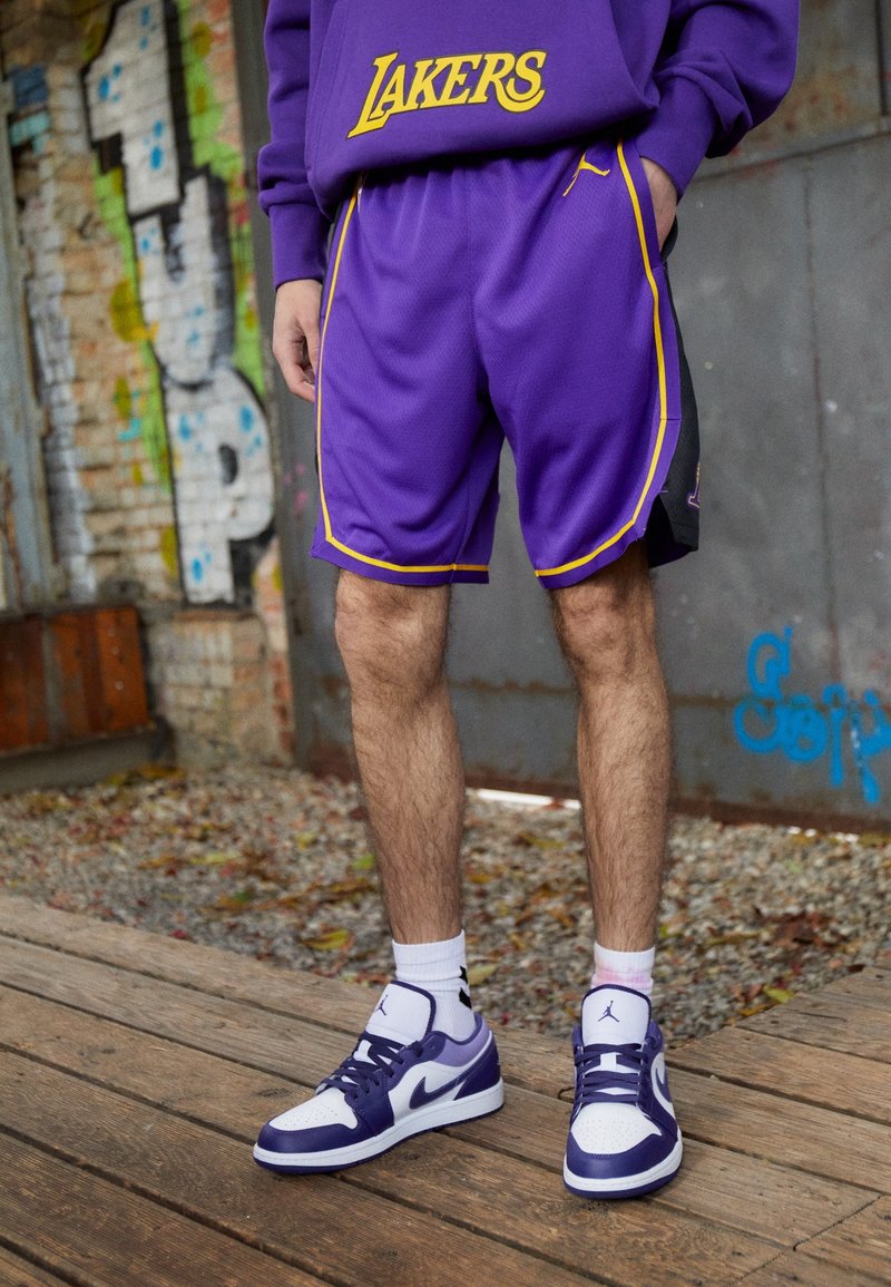 Violet basketballehousut keltaisilla yksityiskohdilla ja yhteensopiva violetin värinen Lakers-huppari. Valkoiset ja violetit lenkkarit täydentävät asun.