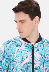 Giacca bomber leggera con uno sfondo rosa e grandi motivi di foglie tropicali blu, colletto navy e zip frontale con tirante ad anello.