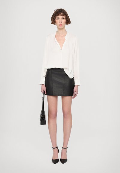 Femme aux cheveux courts et bouclés porte un chemisier blanc, une mini-jupe en cuir noire, des talons noirs à bride cheville et tient un sac à main noir.