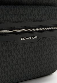 Krupan kadar crne torbe Michael Kors koja prikazuje prepoznatljiv "MK" uzorak, prednji džep sa zatvaračem u srebrnoj boji i srebrnu oznaku branda.