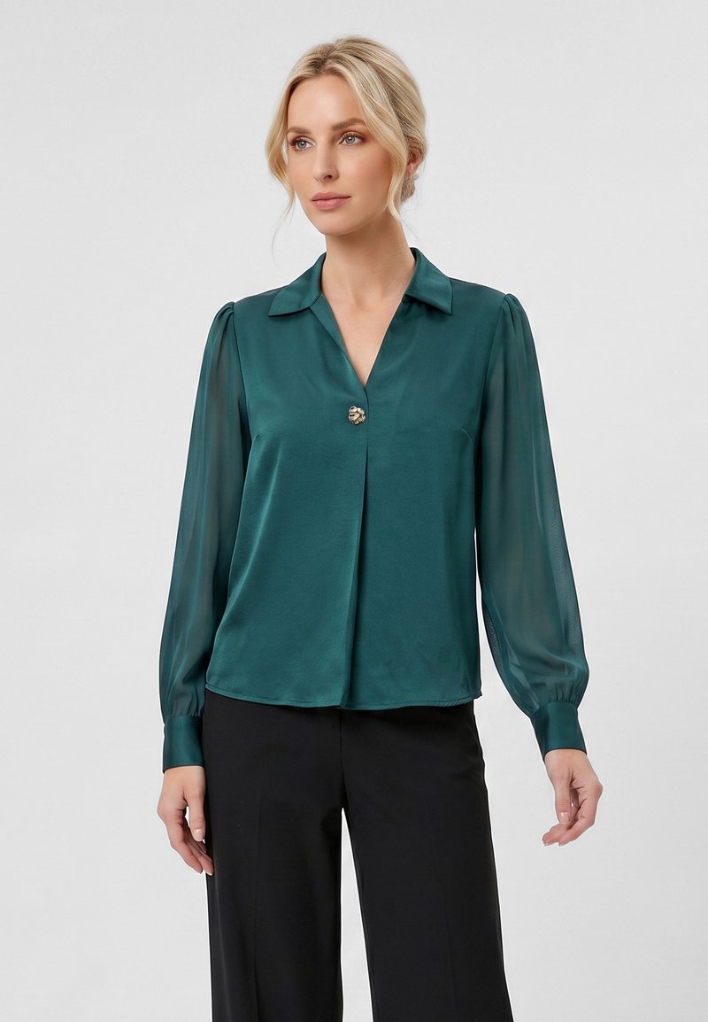 Femme blonde portant un chemisier en satin vert foncé avec de longues manches transparentes et un bouton décoratif unique, associé à un pantalon noir.