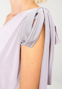 Ljuslila topp med mjuk textur, draperad design och veckade ärmar med knytband vid axeln för extra detalj.