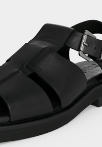 Μαύρο δερμάτινο σανδάλι με φαρδιές λωρίδες, ασημένια αγκράφα και χαμηλό χοντρό τακούνι σε ανοιχτό φόντο.